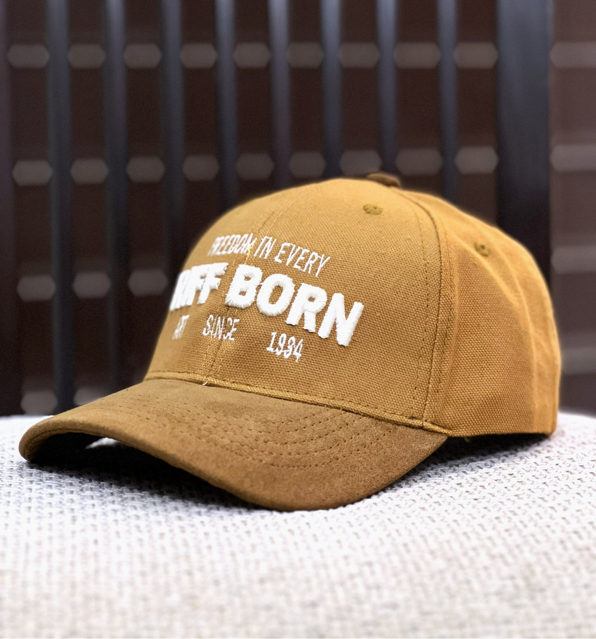 A mustard yellow baseball cap with white embroidered text reading 'RETURN IT EVERY OFF BORN' and 'SINCE 1984', resting on a textured light gray surface against a dark brown slatted background.