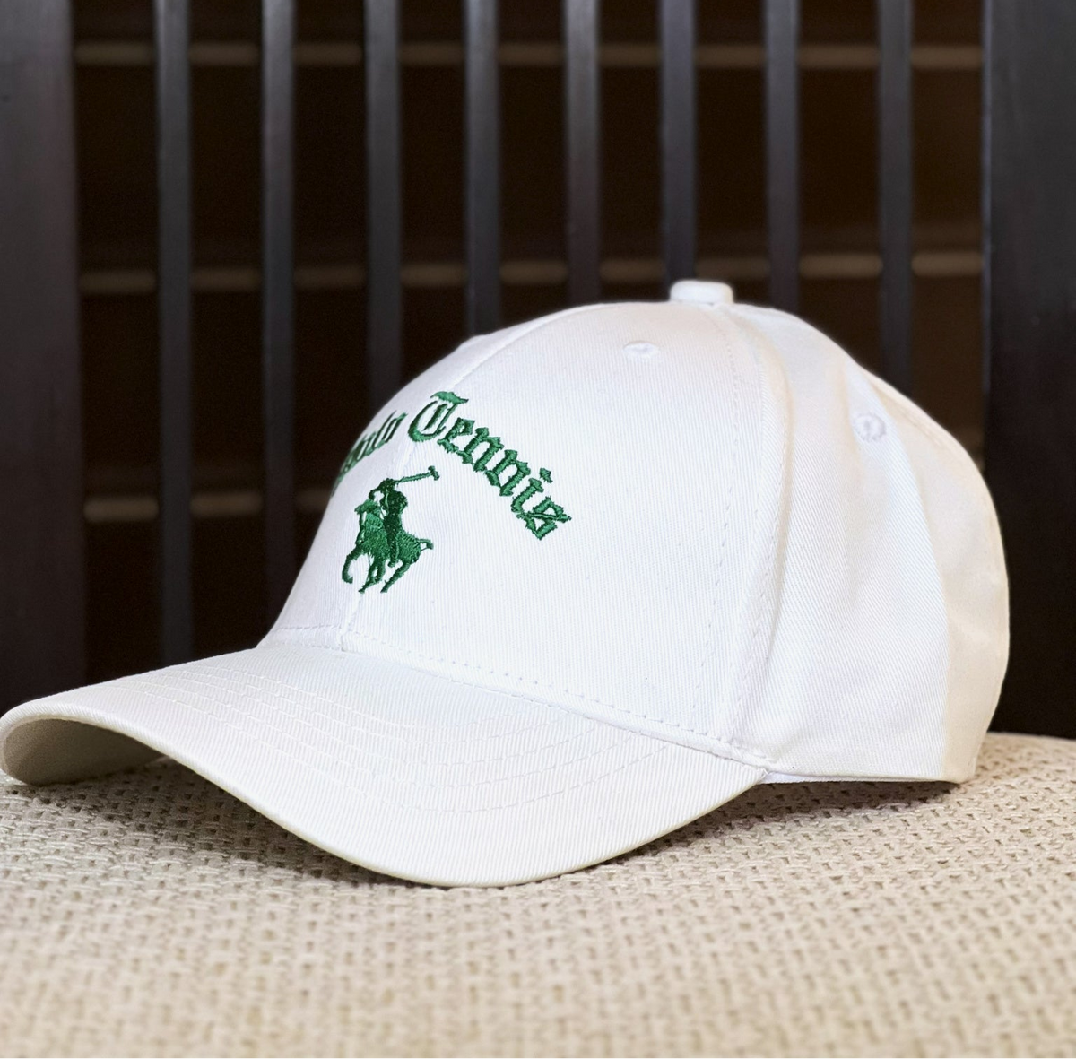 A white baseball cap with green embroidered text reading 'Polo Grounds' and the polo player logo, resting on a textured beige surface in front of a dark wooden slatted background.