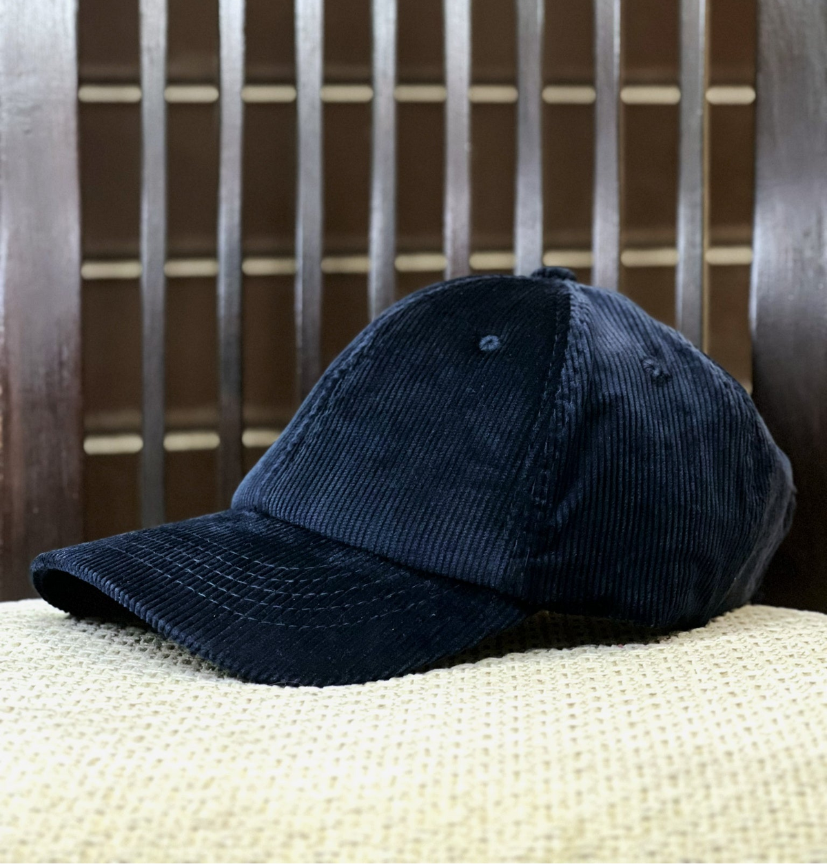 A dark navy blue corduroy baseball cap resting on a textured cream fabric surface in front of a dark brown wooden slatted background.